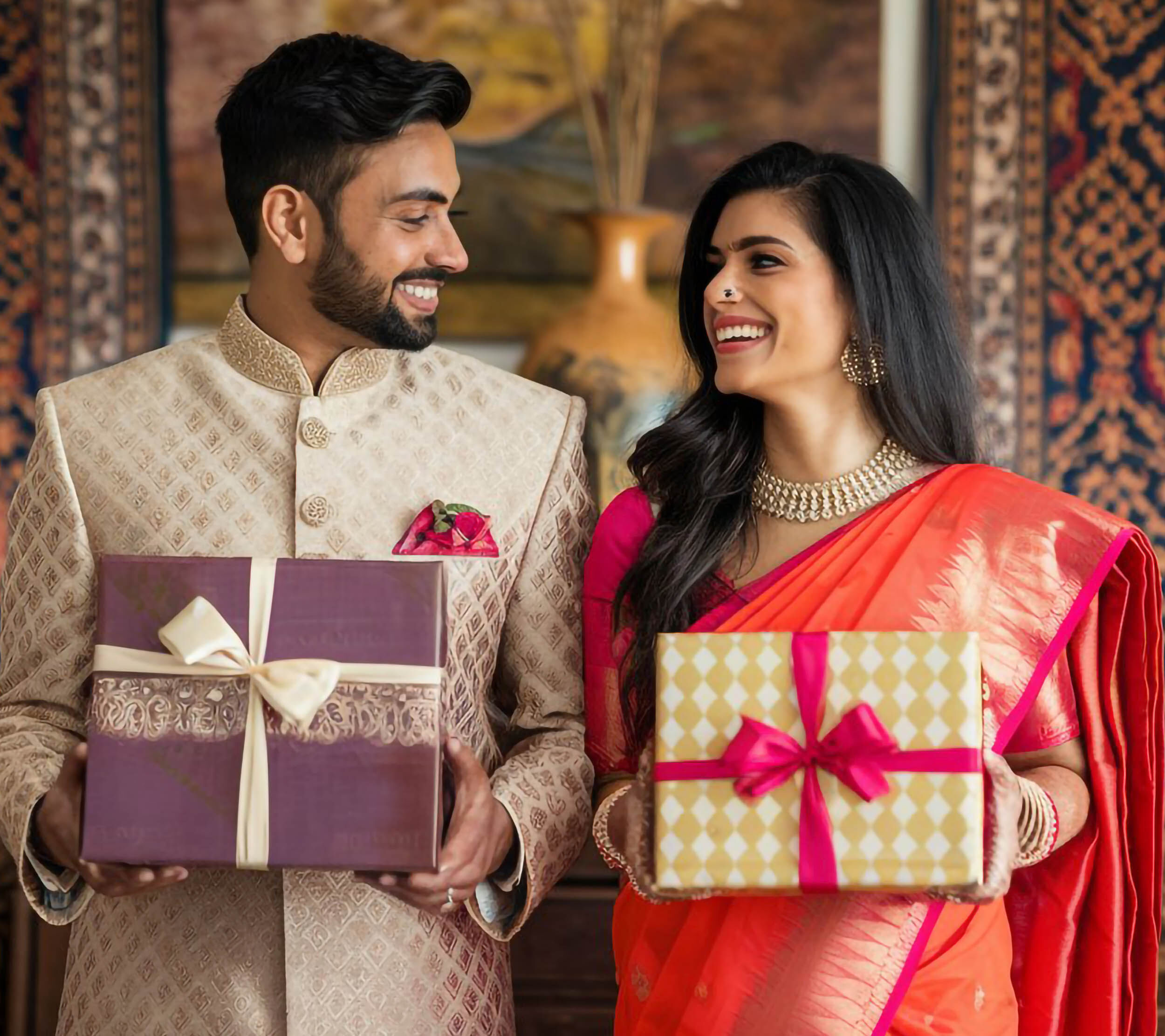 Groom in a beige sherwani and bride in an orange sari holding wrapped wedding gifts